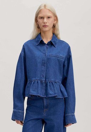 Femme aux longs cheveux blonds portant une veste en jean bleu foncé oversize avec un ourlet peplum et un jean taille haute assorti sur un fond uni.