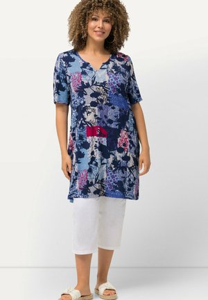 Blaue gemusterte Tunika mit V-Ausschnitt, kurzen Ärmeln und floralen sowie geometrischen Designs. Kombiniert mit weißen Caprihosen und Sandalen.