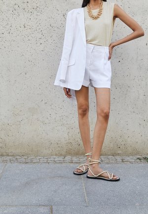 Kobieta w białych szortach, beżowej bluzce bez rękawów, białej marynarce narzuconej na ramię, masywnym złotym naszyjniku oraz beżowych, płaskich sandałach na paski.