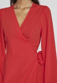 Červené zavinovacie šaty s dlhými, vypchatými rukávmi a výstrihom do V, vyrobené z ľahkej tkaniny. Majú pásový detail s uväzovaním a jemnú textúru.