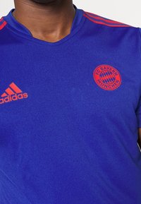 Modré sportovní tričko s kulatým výstřihem, krátkými rukávy, červenými detaily na ramenou a logem FC Bayern München na levé straně hrudi.