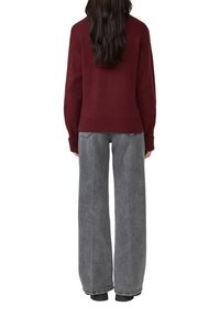 Vrouw met lang zwart haar, gekleed in een bordeauxrode trui en grijze wijde jeans, staande met de rug naar de camera tegen een witte achtergrond.