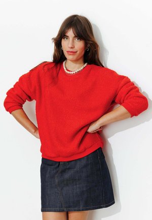 Femme portant un pull rouge texturé, un collier de perles et une jupe en jean foncé, debout les mains sur les hanches sur fond blanc.