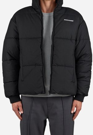 Homme portant une doudoune noire avec le logo "PEGADOR", un pull gris en dessous et un pantalon gris foncé, debout avec les mains détendues le long du corps.