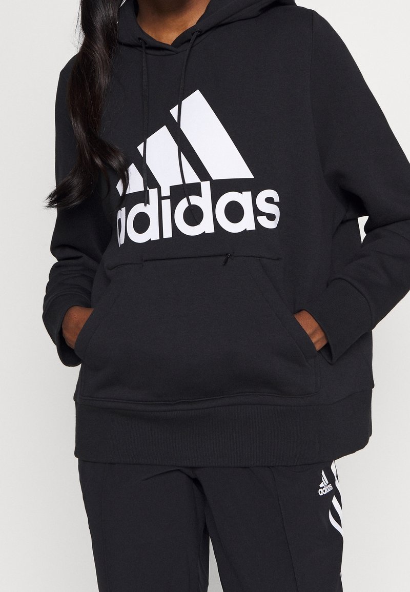 Černá mikina s kapucí vyrobená z bavlněné směsi, s velkým bílým logem Adidas na přední straně a klokankovou kapsou. Měkká textura.