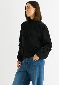Black knitted sweater with a high neck and puffed sleeves, featuring textured heart shapes created from pom-poms across the front.
