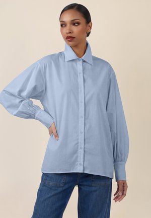 Woman with pulled-back hair wearing a light blue oversized button-up shirt and dark blue jeans, posing with one hand on hip.