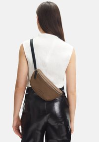 Kvindelig med langt brunt hår iført en hvid ærmeløs top, sorte læderbukser og en brun crossbody taske med en sort rem.