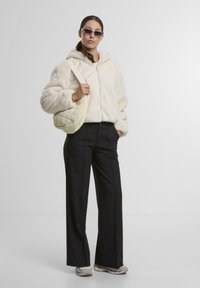 Faux-Fur-Jacke in Creme mit Reißverschluss, schwarze weite Hose, cremefarbene gesteppte Schultertasche und graue Sneaker. Einfaches Design mit Kapuze.