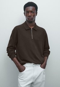 Junger Mann mit Händen in den Taschen trägt ein dunkelbraunes langärmliges Strickoberteil mit Frontreißverschluss und weiße Hose, steht vor einer schlichten hellen Wand.