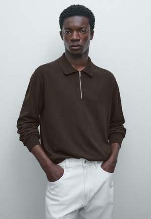 Young man with hands in pockets wears dark brown long-sleeve knit top with front zipper and white pants, standing against plain light wall.