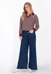Maglione a righe rosse e bianche con maniche lunghe, scollo a V e colletto a contrasto, abbinato a jeans a gamba larga in denim scuro e sneakers bianche.