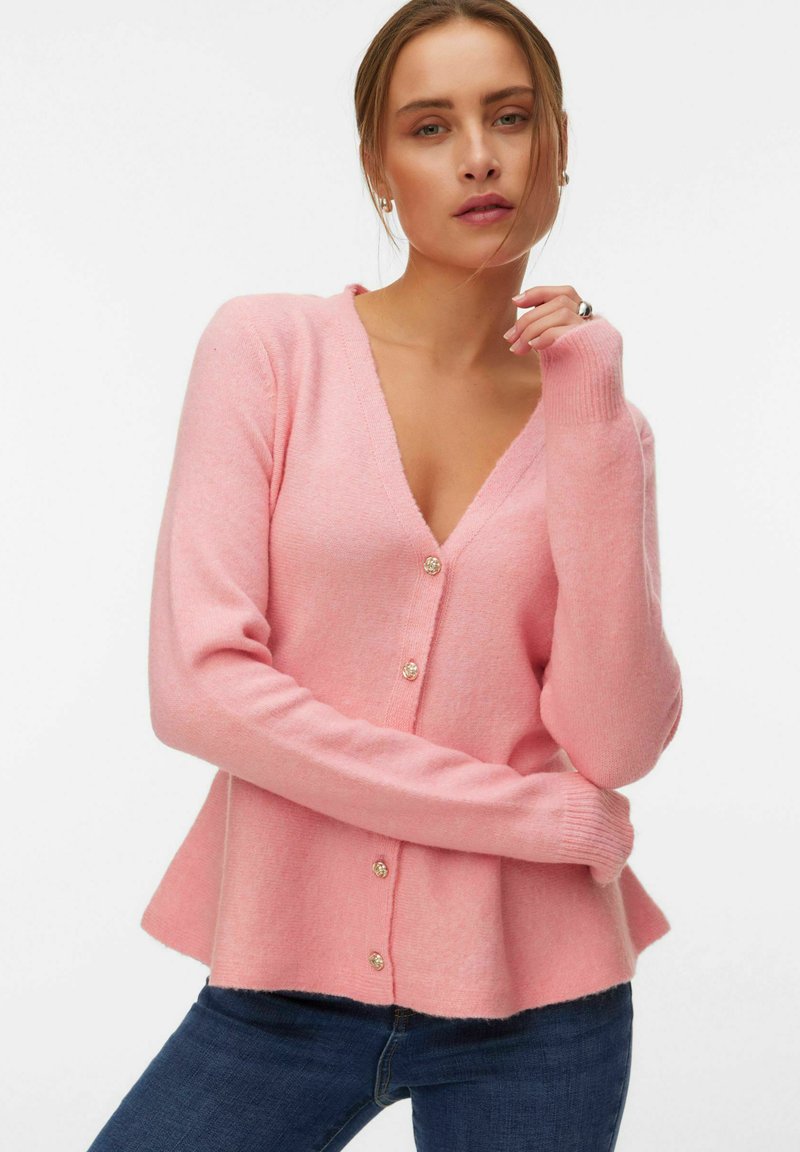 Femme portant un cardigan boutonné rose clair à ourlet évasé et un jean bleu foncé, posant avec un bras croisé et la main près du visage.