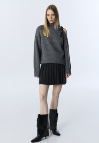 Maglione grigio a maglia con collo alto e aperture sulle spalle; abbinato a una gonna nera plissettata e stivali alti lucidi neri.