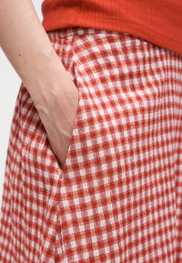 Χέρι που ακουμπά μέσα στην τσέπη μιας κόκκινης και άσπρης καρό φούστας που φοριέται με κόκκινο τοπ, δείχνοντας την υφή του υφάσματος και τη λεπτομέρεια της τσέπης.