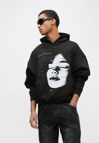 Črna jopa s kapuco z velikim tiskanim obrazom, napisom nad njim in sproščenim krojem. Oblečena z temnimi jeans hlačami in sončnimi očali.