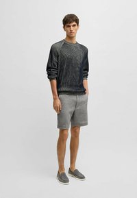 Dunkler, strukturierten Pullover mit einem gerippten Rundhalsausschnitt, kombiniert mit hellgrauen Shorts. Das Modell trägt graue Slipper. Schlichtes, modernes Design.