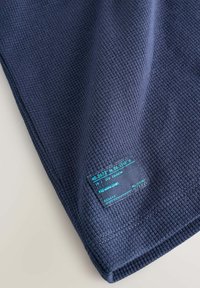 Detail tmavomodrej tkaniny s vaflovým vzorom so malou čiernou záplatou, na ktorej je svetlomodrý text a súradnice.