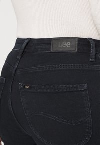Jeans de mezclilla oscura con una superficie texturizada, que presentan una etiqueta de cuero negro con la marca "Lee" y un pequeño logo dorado en el bolsillo.