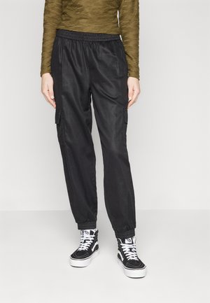 Zwarte cargo-broek met elastische taille en boorden, met twee zijzakken. G gedragen met zwart-witte hoge sneakers. Getextureerde olijfgroene top.