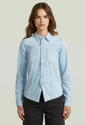 Woman with wavy brown hair wearing a light blue button-up denim shirt and black pants, standing against a plain background.