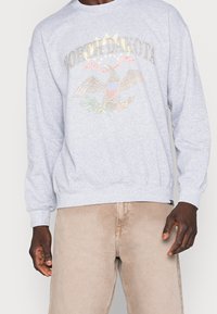 Sudadera gris con un gráfico de un águila y el texto "NORTH DAKOTA". Mangas largas, puños y dobladillo de canalé con un corte relajado. Fondo liso.