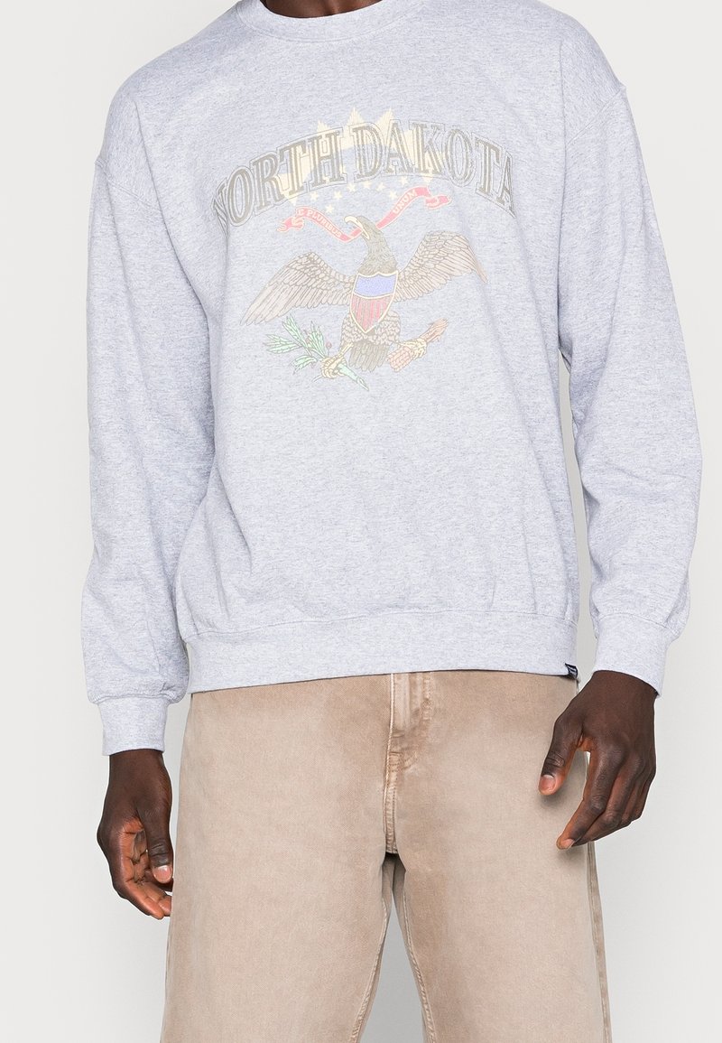 Sudadera gris con un gráfico de un águila y el texto "NORTH DAKOTA". Mangas largas, puños y dobladillo de canalé con un corte relajado. Fondo liso.
