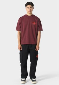 Nuori mies seisoo, päällään maroon lyhythihainen paita, jossa on punainen graafinen kuvio, sekä mustat cargo-housut, joissa on vastaava punainen graafinen kuvio taskussa.