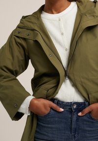 Olive green lightweight jacket with a zip front, adjustable hood, and snaps; worn over a white ribbed sweater and dark denim jeans.