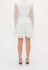 Witte jurk met een bloemenpatroon, lange doorschijnende mouwen, geruite lagen en een getailleerde taille, gecombineerd met zwarte en witte hoge hakken met bandjes.