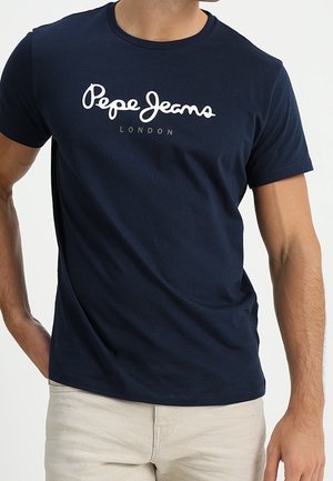 Mand iført en mørkeblå t-shirt med hvidt "Pepe Jeans London"-logo, kombineret med lyse beige bukser, stående med den ene hånd på hoften.