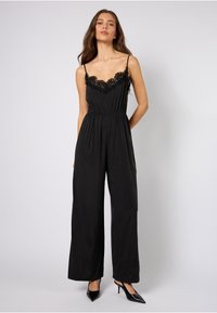 Sort jumpsuit med blonderkant ved utringningen, spaghettistropper, elastisk midje og vide ben. Brukt med sorte høye hæler.