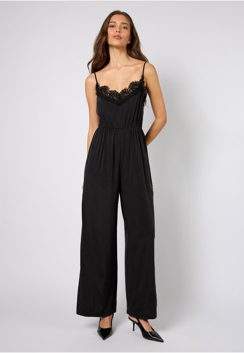 Sort jumpsuit med blonderkant ved utringningen, spaghettistropper, elastisk midje og vide ben. Brukt med sorte høye hæler.