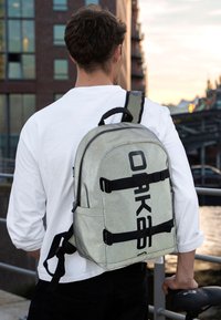 Svetlošedý ruksak s čiernym textom, so zipsovými priehradkami, nastaviteľnými popruhmi a textúrovaným povrchom, nosený na chrbte osoby.