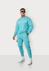 Türkises Sweatshirt und Jogginganzug-Set mit weißem Logo. Weicher Stoff, lockere Passform, gerippte Bündchen und Cargo-Taschen. Kombiniert mit weißen Turnschuhen.