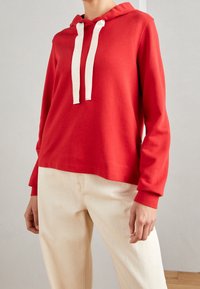 Sudadera roja con capucha, confeccionada en tejido suave, con cordones blancos, mangas largas y un corte holgado. Combinada con pantalones de tonos claros.