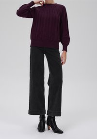 Dunkellila gestrickter Pullover mit strukturierten Mustern, weiten Ärmeln und geripptem Saum, kombiniert mit hochgeschnittenen schwarzen weiten Jeans und schwarzen Knöchelstiefeln.