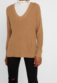 Beżowy sweter z dekoltem w serek o prążkowanej fakturze i długich rękawach, zestawiony z czarnymi, podartymi dżinsami. Sweter ma luźny krój i boczne rozcięcia.