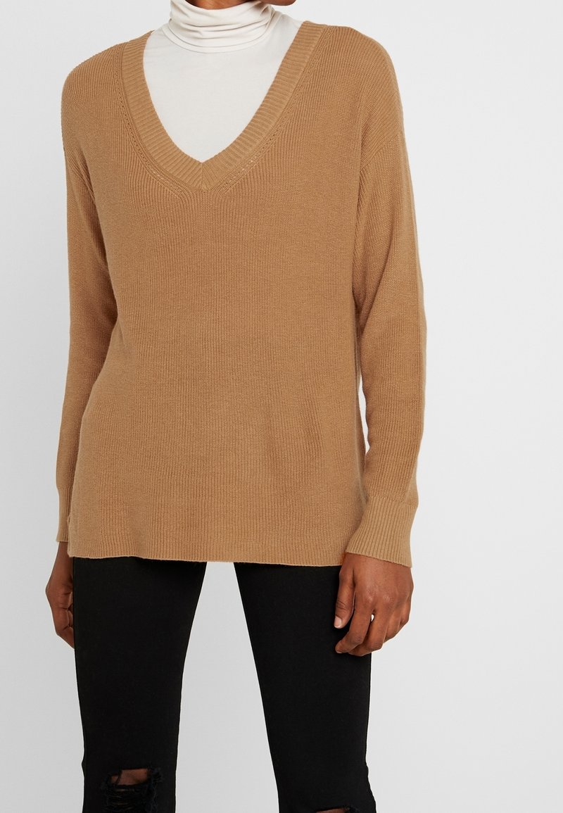 Beżowy sweter z dekoltem w serek o prążkowanej fakturze i długich rękawach, zestawiony z czarnymi, podartymi dżinsami. Sweter ma luźny krój i boczne rozcięcia.