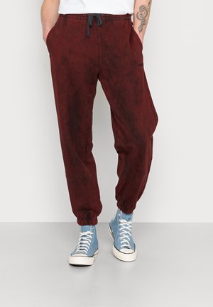 Personne portant un pantalon de jogging rouge foncé avec des chevilles élastiques, les mains dans les poches, une chemise blanche et des baskets montantes bleu clair.