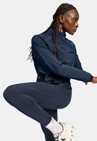 Marineblauer Windbreaker mit hohem Kragen und Reißverschluss vorne, kombiniert mit dunklen Leggings und klobigen weißen Turnschuhen. Verfügt über reflektierende Akzente.