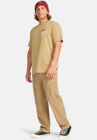 Beige bomull t-skjorte med grafisk trykk, sammen med brune cordfløyelsbukser. Sorte joggesko med hvite såler fullfører antrekket.