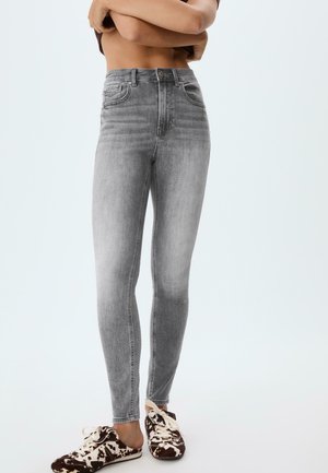 Frau trägt hoch taillierte graue Skinny-Jeans und schwarz-weiße Sneakers mit Muster, steht mit verschränkten Armen über dem bloßen Oberkörper.