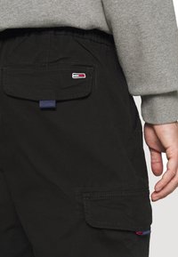 Černé nákladní kalhoty s dvojitými zadními kapsami, malým vyšívaným logem a elastickým pasem. Látka vypadá odolně s hladkou strukturou.