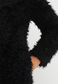 Sort, lodden frakke med lange ærmer, der har et struktureret, fyldigt stof. Materialet virker blødt og har en pjusket finish.