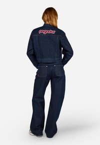 Giubbotto di jeans con logo "pegador" rosso sul retro, abbinato a jeans a gamba larga, entrambi in lavaggio blu scuro, con cuciture visibili e tasche.