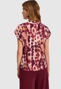 Kobieta odwrócona plecami, ubrana w bluzkę z krótkim rękawem w abstrakcyjny wzór w czerwieni, różu i fioletu, w połączeniu z burgundowymi spodniami na jednolitym tle.