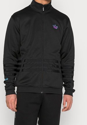 Mężczyzna ubrany w czarną kurtkę na zamek błyskawiczny z logo Adidas w kolorze fioletowo-niebieskim na klatce piersiowej, czarne paski na rękawach i w pasie oraz dopasowane czarne spodnie.