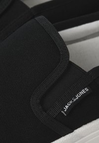 Fekete slip-on cipő texturált anyagból, eloxált oldalakkal és gumitalppal, fehér kerettel. Jack & Jones logó látható.