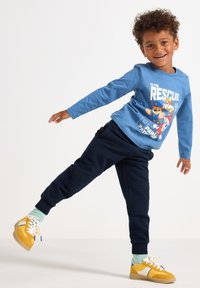 Blaues Langarmshirt mit einem Cartoon-Graphic, kombiniert mit dunkelblauen Jogginghosen mit strukturierten Einsätzen. Gelbe Sneakers mit weißen Akzenten.